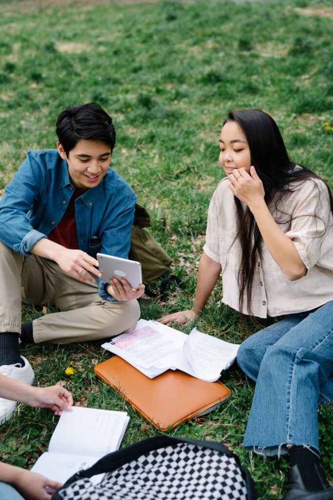 Group of students studying outdoors on grass, using a tablet for research.